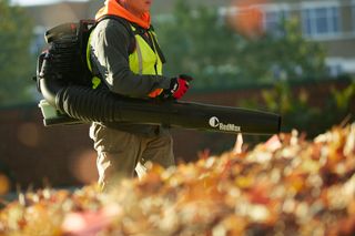 A person using a RedMax leaf blower to clear fallen leaves in a park setting.