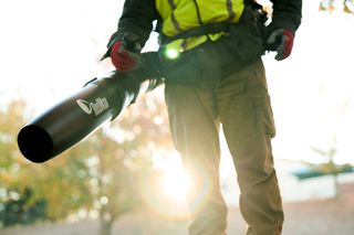 A person using a leaf blower in a park setting, with sunlight filtering through trees.