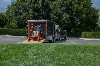 Two people unloading a ride-on mower from a trailer in a parking lot.